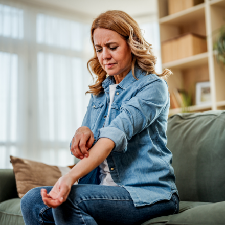 Woman sitting on a couch scratching her itchy forearm, wincing in discomfort