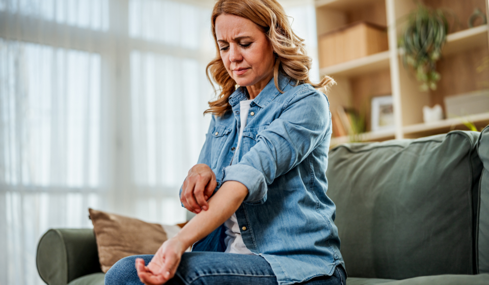 Woman sitting on a couch scratching her itchy forearm, wincing in discomfort